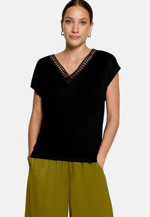 Femme portant un haut noir à manches courtes avec un décolleté en V en dentelle et un pantalon ample vert olive, debout les mains dans les poches.