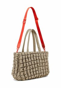 Sac fourre-tout en beige matelassé avec une texture duveteuse, doté de deux poignées courtes et d'une sangle rouge amovible et ajustable. Design rectangulaire.