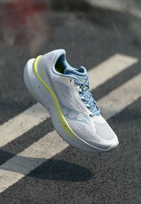 Lys blå joggesko med mesh-overdel, grå detaljer og en gul stripe på siden. Har en dempet såle og teksturerte lisser.