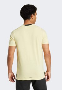 Camiseta amarilla de manga corta con dobladillo recto, acento en el cuello negro y costuras planas. La vista trasera muestra la textura suave de la tela y la marca en la parte inferior.