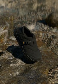 Pantof cu talpă groasă, de exterior, de culoare neagr�ă, cu închidere laterală, așezat pe pietre texturate, în teren natural, stâncos.