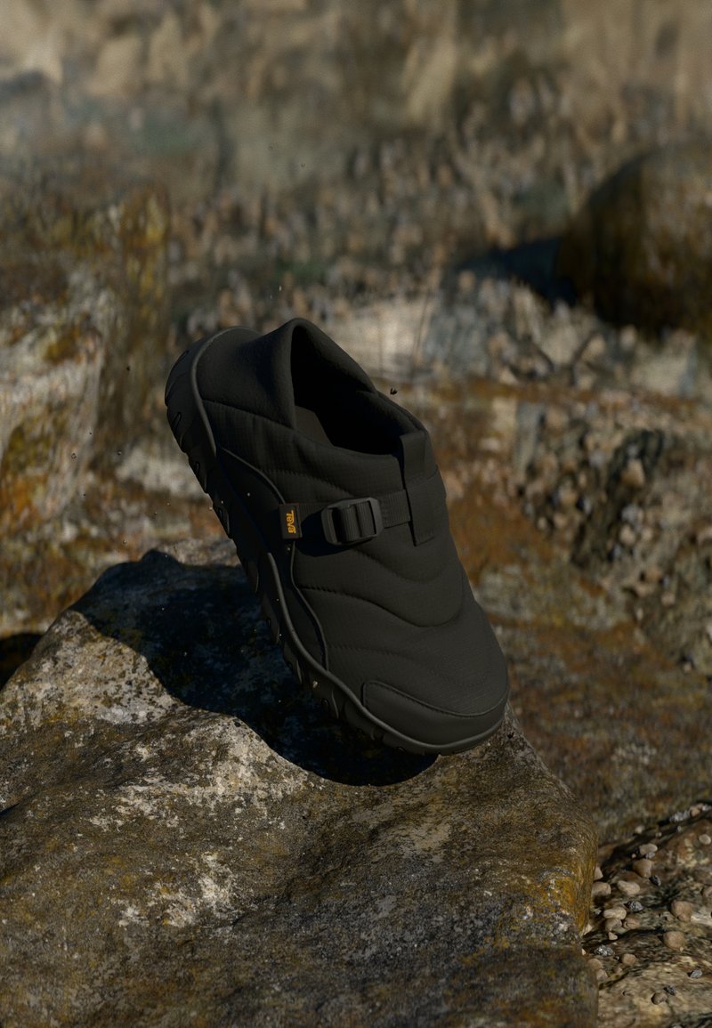 Sort slip-on udendørssko med tyk sål og sidelås, hvilende på strukturerede klipper i naturligt, klippeagtigt terræn.