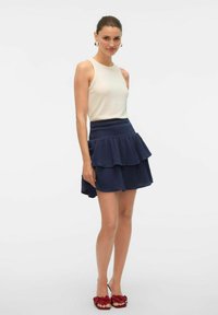 Krämfärgad linne topp kombinerad med en mörkblå lager på lager volangkjol. Outfittet har en texturerad tyg, och modellen bär röda blommiga slip-on-skor.