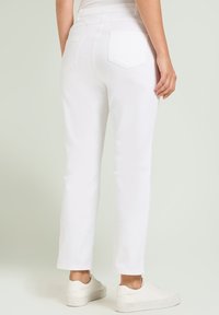 Persoon in witte rechte broeken en witte sneakers, staand tegen een lichtgrijze achtergrond, gezien vanachter.