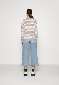 Nainen seisoo selkä kameraan päin, päällään vaaleanbeige ja valkoinen raidallinen pitkähiha shirt, vaaleansiniset leveät 7/8-housut, mustat sukat ja valkoiset kengät.
