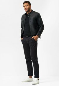 Zwarte leren jas met een staande kraag, gecombineerd met een zwarte trui en donkere broeken. Witte sneakers maken de outfit af.
