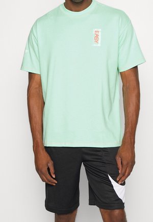 Mężczyzna ubrany w pastelowo zielony t-shirt z napisem "EASY" na piersi, połączony z czarnymi szortami z białym, skośnym paskiem na lewej nodze.