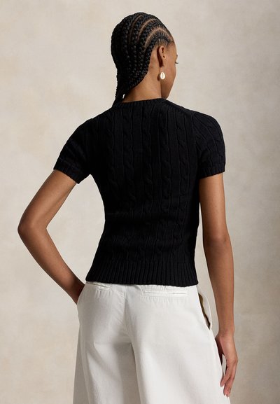 Mujer con el cabello trenzado, vestida con un suéter negro de punto con mangas cortas y pantalones blancos, de pie con una mano en la cadera, de espaldas.