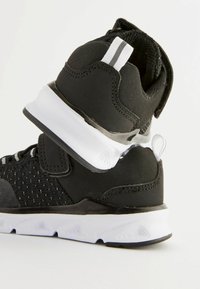 Chaussures de sport noires avec tige en maille, amorti léger, semelle blanche et boucle à l'arrière. Présente des rayures argentées et des détails texturés.