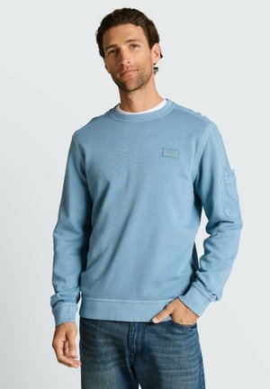 Mężczyzna z krótkimi kręconymi włosami, ubrany w jasnoniebieską bluzę z okrągłym dekoltem i niebieskie dżinsy, stoi z jedną ręką w kieszeni na jednolitym tle.