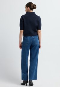 Marineblaues kurzärmliges Strickpullover mit umgeschlagenem Kragen, kombiniert mit hoch taillierten blauen Jeans. Rückansicht, die eine schlanke Passform und strukturierten Stoff zeigt.