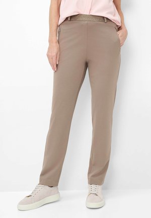 Donna che indossa pantaloni taupe a gamba dritta con tasche laterali con cerniera e scarpe da ginnastica beige, in piedi su sfondo bianco.