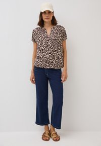 Blouse à manches courtes avec motif léopard marron et beige, associée à un jean foncé à jambes larges et des sandales à lanières beiges ; casquette légère sur la tête.