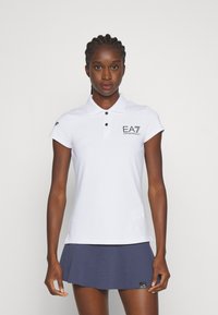 Polo bianco con colletto, maniche corte e logo EA7 nero. Abbinato a una gonna a righe blu scuro. Tessuto morbido e design aderente.