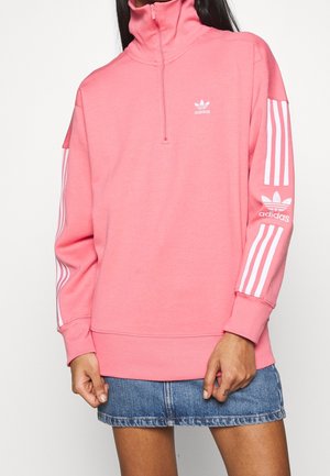 Žena nosící růžový pulovr Adidas s bílými pruhy a logem na rukávech, doplněný modrou džínovou minisukní.