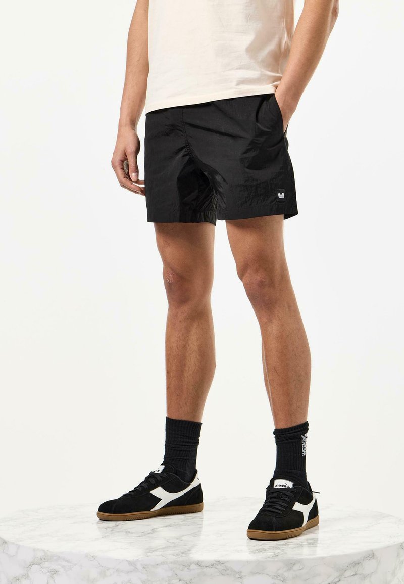 Schwarze Shorts aus leichtem Material, mit seitlichen Taschen, lockerer Passform und einem kleinen Logodetail. Kombiniert mit schwarzen Schuhen und Socken.