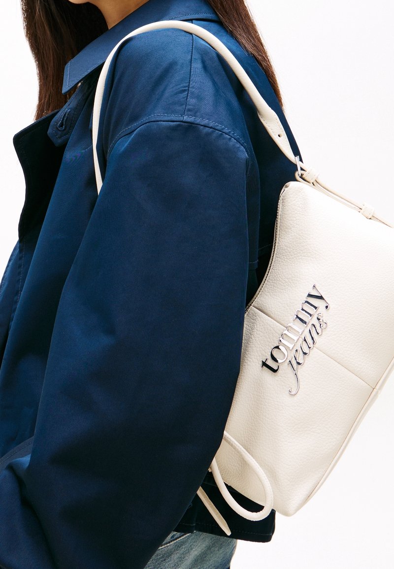 Une femme portant une veste bleu foncé porte un sac à bandoulière blanc Tommy Jeans avec un lettrage de logo argenté.