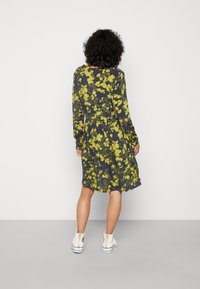 Vestido floral com mangas longas, fundo azul-marinho, flores amarelas brilhantes, cintura ajustada e saia rodada que termina acima do joelho. Usado com ténis brancos.