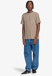 Beżowa bawełniana koszulka z kieszonką na piersi, w połączeniu z luźnymi niebieskimi dżinsami. Czarne trampki z białymi akcentami i sznurowadłami.