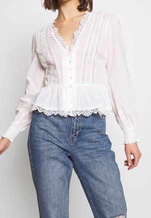 Kobieta w białej, długorękawowej bluzce z koronkową lamówką i guzikami z przodu, połączona z wysokim stanem niebieskich dżinsów.