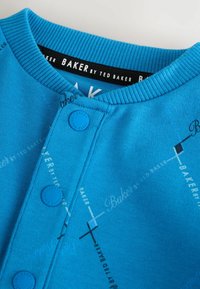 Blaues Baumwolloberteil mit Rundhalsausschnitt, auffälligen Knopfdetails und subtilem, sich wiederholendem Textmuster in Weiß und Schwarz.