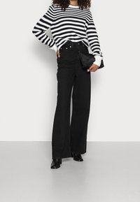 Czarne dżinsy z wysokim stanem i szerokimi nogawkami w połączeniu z czarno-białym swetrem w paski, trzymane w czarnej torebce kopertowej. Gładki materiał, casualowy fason.