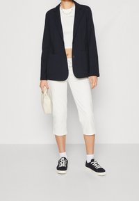 Námořnický blazer se dvěma předními kapsami, bílý cropped top, bílé cropped kalhoty a tmavé tenisky s bílou podrážkou a tkaničkami, držící krémovou tašku.