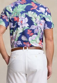 Camicia a maniche corte con stampa floreale in colori viola, verde e rosa; abbinata a pantaloni bianchi e una cintura marrone; tessuto a trama liscia.