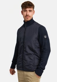 Navy puffer jacket with a high collar and knit sleeves. Features a zip front and logo patch on the left sleeve. Lightweight and structured design.