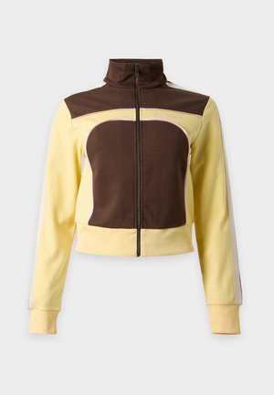 Joven mujer con chaqueta amarilla y marrón, camiseta negra, vaqueros anchos azules, sosteniendo un pequeño bolso marrón, de pie frente a un fondo blanco liso.
