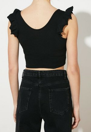 Person trägt ein ärmelloses schwarzes Crop-Top mit Rüschen an den Schultern und hoch taillierte schwarze Denim-Jeans, von hinten gezeigt.