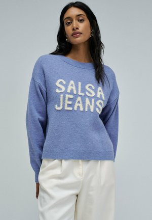 Suéter azul claro con la inscripción "SALSA JEANS" en letras texturizadas color crema. Mangas largas, corte holgado y dobladillo acanalado. Combinado con pantalones color crema.
