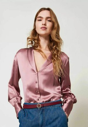 Blusa di raso rosa con scollo a V, maniche lunghe e chiusura con bottoni. Abbinata a jeans blu a vita alta e una larga cintura bordeaux.