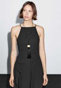 Černé šaty s halter výstřihem a volným stříhem, s dlouhým černým náhrdelníkem s třásněmi a hladkým, matným povrchem.
