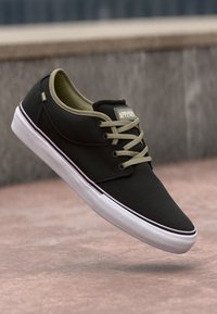 Black canvas sneaker with olive green laces, white midsole, and subtle branding on the tongue. Features a low-profile design and rounded toe.