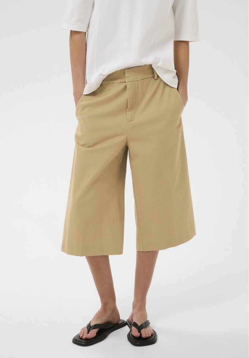 Personne portant un short beige mi-long, une chemise blanche à manches courtes et des sandales noires à bouts carrés, mains dans les poches.