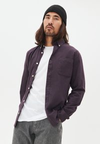 Ung man med långt hår och skägg som bär en svart mössa, en upplåst mörk lila skjorta över en vit t-shirt och grå jeans, med händerna i fickorna.