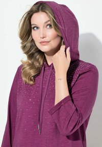 Violetti huppari, jossa on nyöri ja hopeiset koristeet edessä. Kevyt, teksturoitu kangas, pitkät hihat ja rento istuvuus.