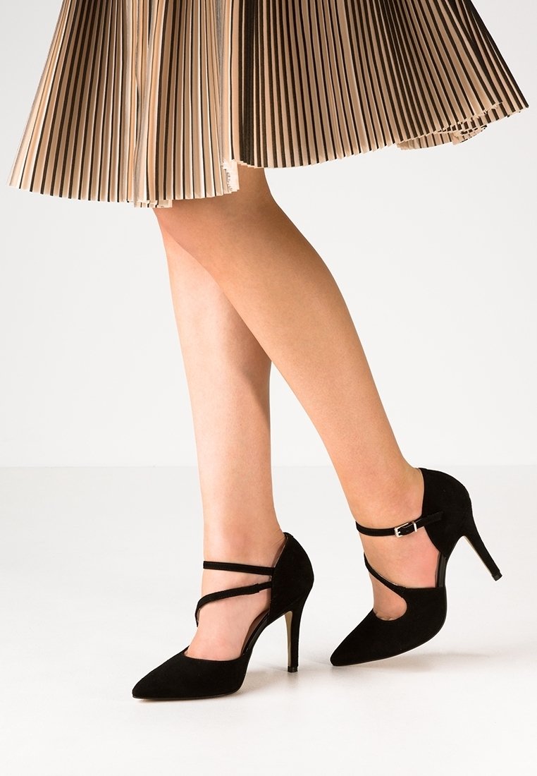 Femme portant une jupe plissée beige et des escarpins en daim noir avec des brides à la cheville, debout sur la pointe des pieds sur un fond blanc uni.