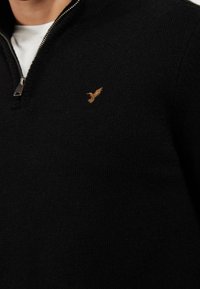 Pull noir en maille avec un col montant et une fermeture éclair, orné d'une petite broderie dorée d'oiseau sur le côté gauche de la poitrine.