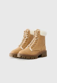 Bottes lacées beige avec un col en fausse fourrure, semelle extérieure robuste et œillets métalliques. Matériau en daim lisse avec des détails de couture contrastants.