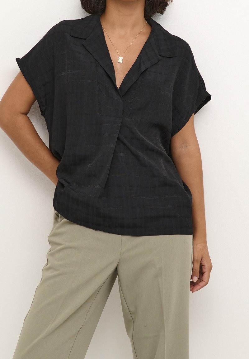 Femme portant un chemisier noir à manches courtes avec un col et un pantalon beige, debout avec une main posée sur la hanche.