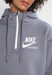 Ljusgrå hoodie i mjukt tyg, med en vit dragkedja, snörning och Nike-logotyp med texten "SPORTSWEAR" i vitt.
