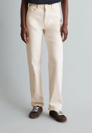 Osoba nosząca kremowe proste dżinsy i ciemnobrązowe sznurowane trampki, stojąca na tle jasnoszarym.