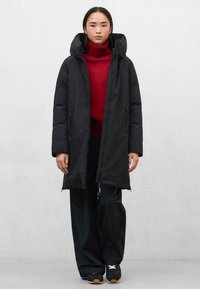 Černý dlouhý prošívaný kabát s kapucí, se zapínáním na přední zip a bočními kapsami, nošený přes červený rolák a široké černé kalhoty.