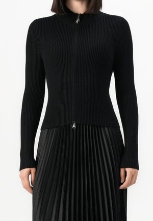 Kobieta ubrana w dopasowany, prążkowany, czarny sweter na zamek w połączeniu z czarną plisowaną spódnicą z wysokim stanem, stojąca na białym tle.