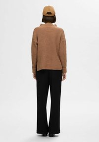Pull marron en tricot avec texture côtelée, vu de dos, associé à un pantalon noir à jambes larges et une casquette de baseball beige.