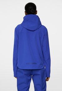 Giacca blu realizzata in tessuto liscio e leggero, con cappuccio, polsini elasticizzati e un logo stampato nella parte bassa della schiena.