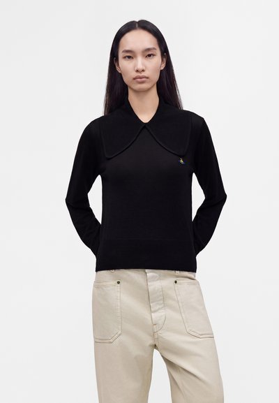 Personne portant un haut noir à manches longues avec un grand col et un pantalon beige, debout les mains dans le dos sur un fond uni.