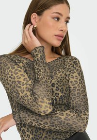 Donna con capelli lunghi che indossa orecchini a cerchio irregolari in oro e una blusa trasparente a maniche lunghe con stampa leopardata, toccandosi l'orecchio e guardando di lato.